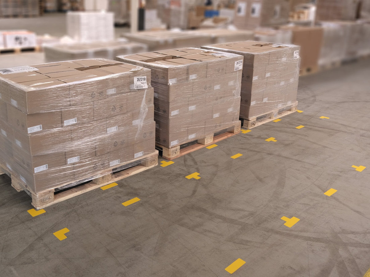 Yellow T-shaped floor markers set out between wrapped pallets in a warehouse