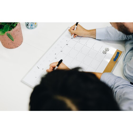 Two people writing on a Quartet combo calendar planner QTCP406508 laid on a table, with blue push pins in the cork strip and round magnets on the grid