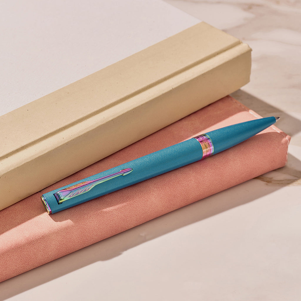 Teal Meridian ballpoint in a styled desk setting, showing the rainbow clip and centre band